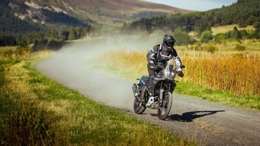 Rendimiento de la llanta MICHELIN Anakee Adventure 2: conducción de motocicleta en un camino de grava con colinas y campos verdes en el fondo.