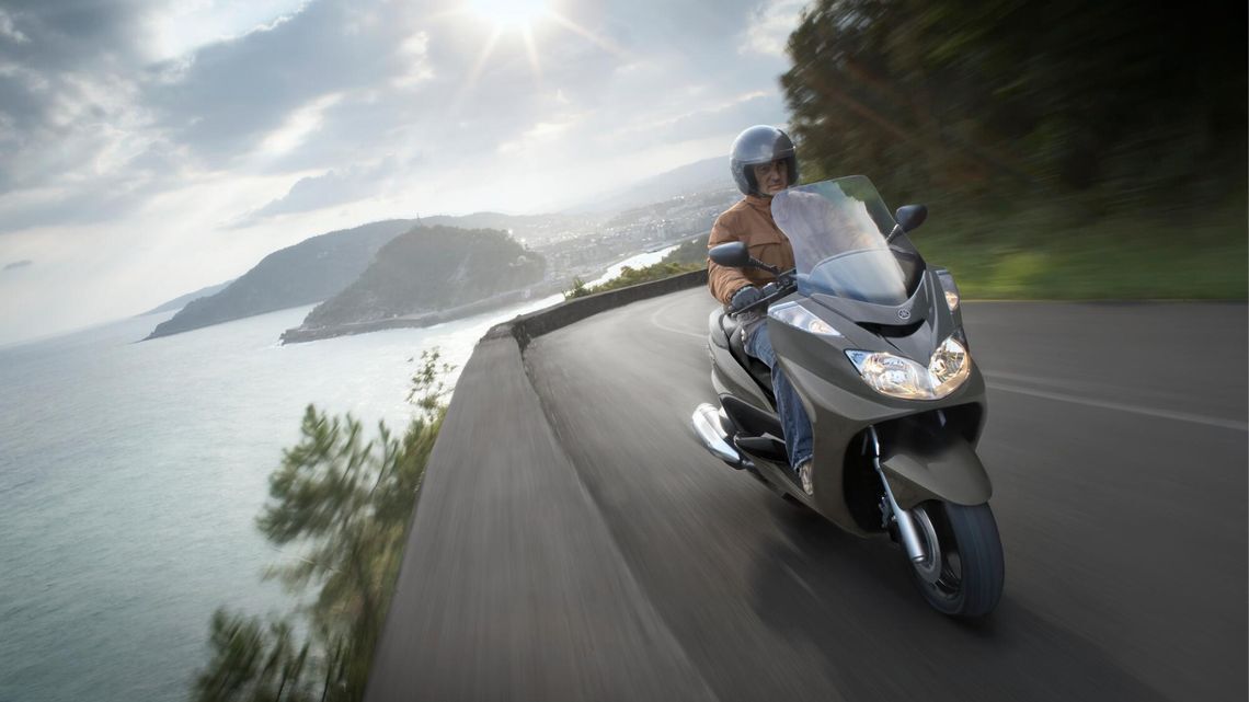 A rider on a motorcycle travels a coastal road, with the ocean and a bright sky creating a picturesque backdrop