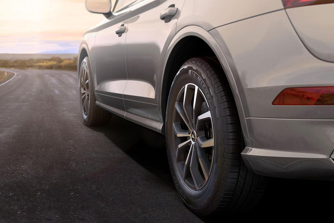 Close-up view of the rear side and wheel of a beige SUV parked on an empty paved road, with a landscape and sky in the background.