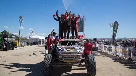 El equipo ganador celebra la victoria sobre el techo de un buggy Baja, rociando con champán a la multitud y la línea de meta