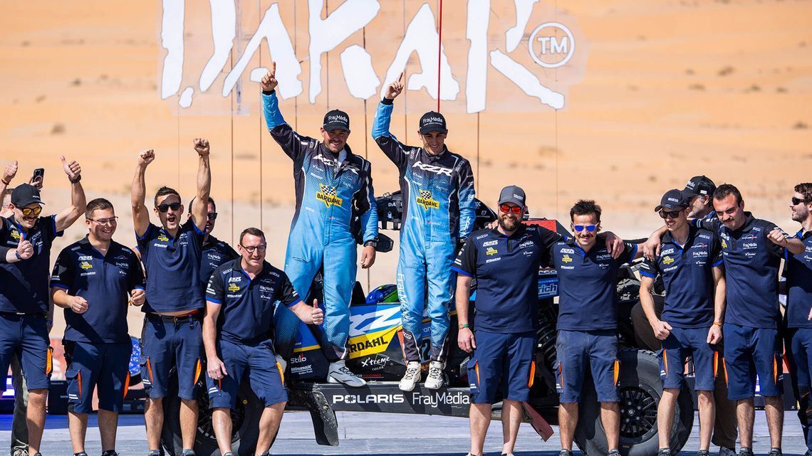 Dakar-rallitiimi juhlii Dakar-logon edessä voittoa niin, että kaksi sinisissä haalareissa olevaa kuljettajaa seisoo ajoneuvon päällä ja heidän ympärillään ovat tiimin jäsenet univormuissaan.