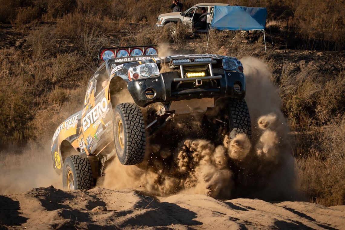 En tävlingslastbil över en sanddyn, rör upp ett moln av damm, under en Baja-tävling