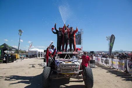 Tävlingsteam firar seger på taket av en Baja-buggy, sprejande publiken och mållinjen med champagne