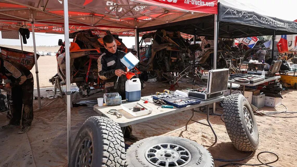 Mekaniker arbetar på terrängfordon under tält i ett serviceområde på Dakar-rallyt, omgiven av reservdäck, verktyg och utrustning