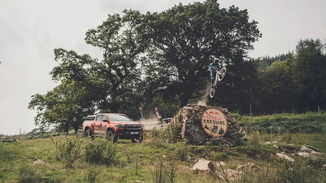 Mountainbikecyklist hoppar över en sten märkt Red Bull Hardline, intill en orange Ford Ranger-pickup, i en naturskön miljö med träd och buskar.