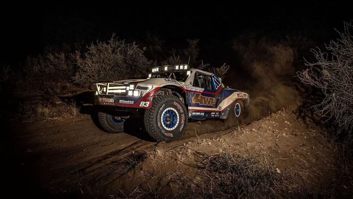 El camión de carreras todoterreno de color rojo, blanco y azul, con el número 83 y el logotipo de BFGoodrich, conduciendo de noche por un terreno polvoriento con los faros encendidos.