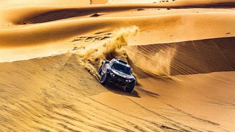 Un vehículo de rally todoterreno cruza una duna de arena en el desierto levantando una nube de polvo