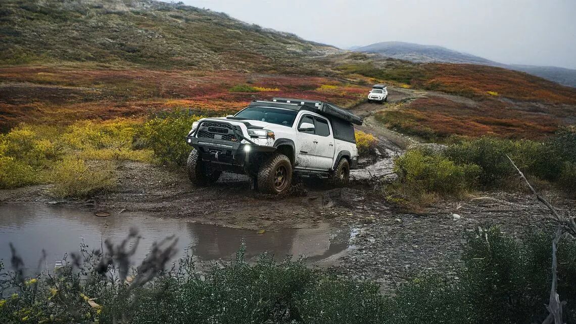 Furgoneta pick up blanca con tienda de campaña en el techo y neumáticos BFGoodrich todoterreno, atravesando terrenos embarrados con charcos y colorida vegetación otoñal