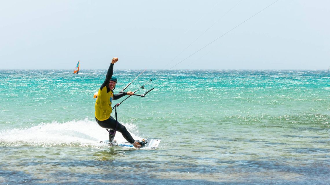 Personne faisant du kitesurf sur une mer turquoise, vêtue d’un t-shirt jaune et d’une combinaison noire, un bras levé dans les airs