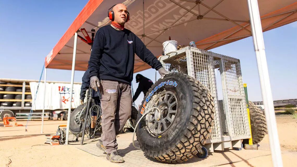 Mekaniker i svart collegetröja och grå byxor pumpar upp ett stort BFGoodrich-terrängdäck i ett tält i öknen, omgiven av verktyg och utrustning