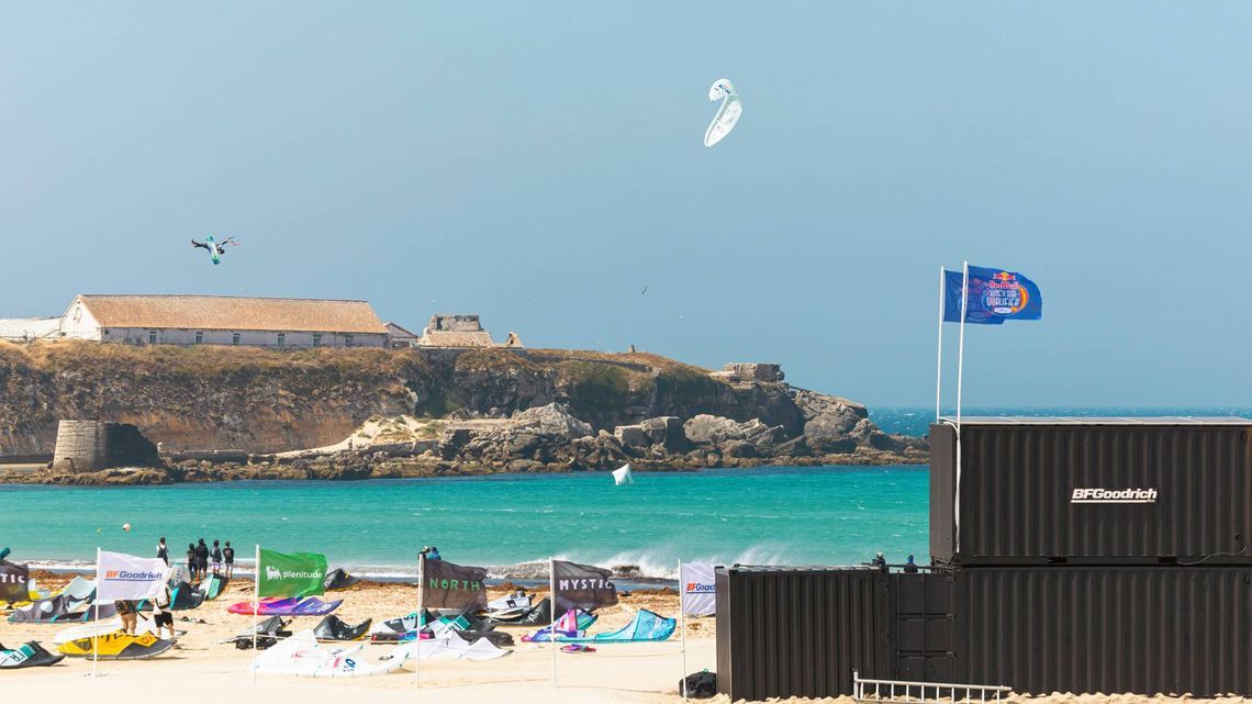 Un kiteboarder s'envole au-dessus de la mer, lors de la compétition Red Bull King of the Air à Tarifa, avec des drapeaux d'événements et des structures temporaires sur la plage au premier plan