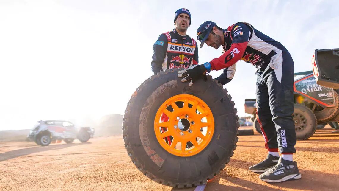 Rallyförare i overaller och kepsar inspekterar ett BFGoodrich orange rim-däck, med rallybilar och utrustning i bakgrunden