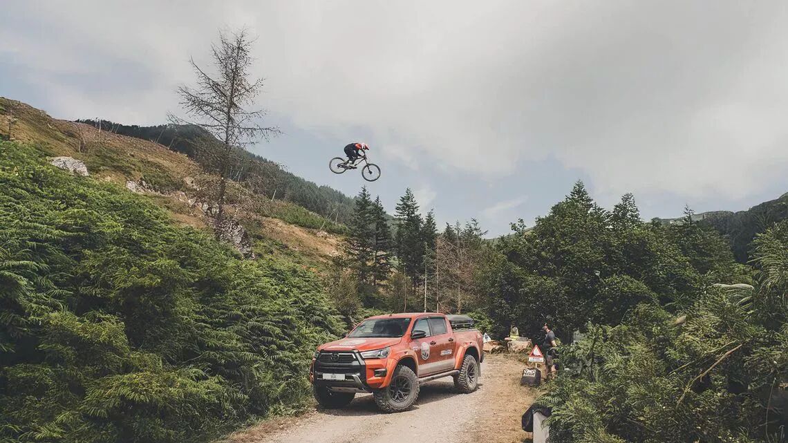 Ciclista de montaña realizando un espectacular salto sobre una furgoneta pick up roja en medio de un camino de tierra en un bosque verde con colinas