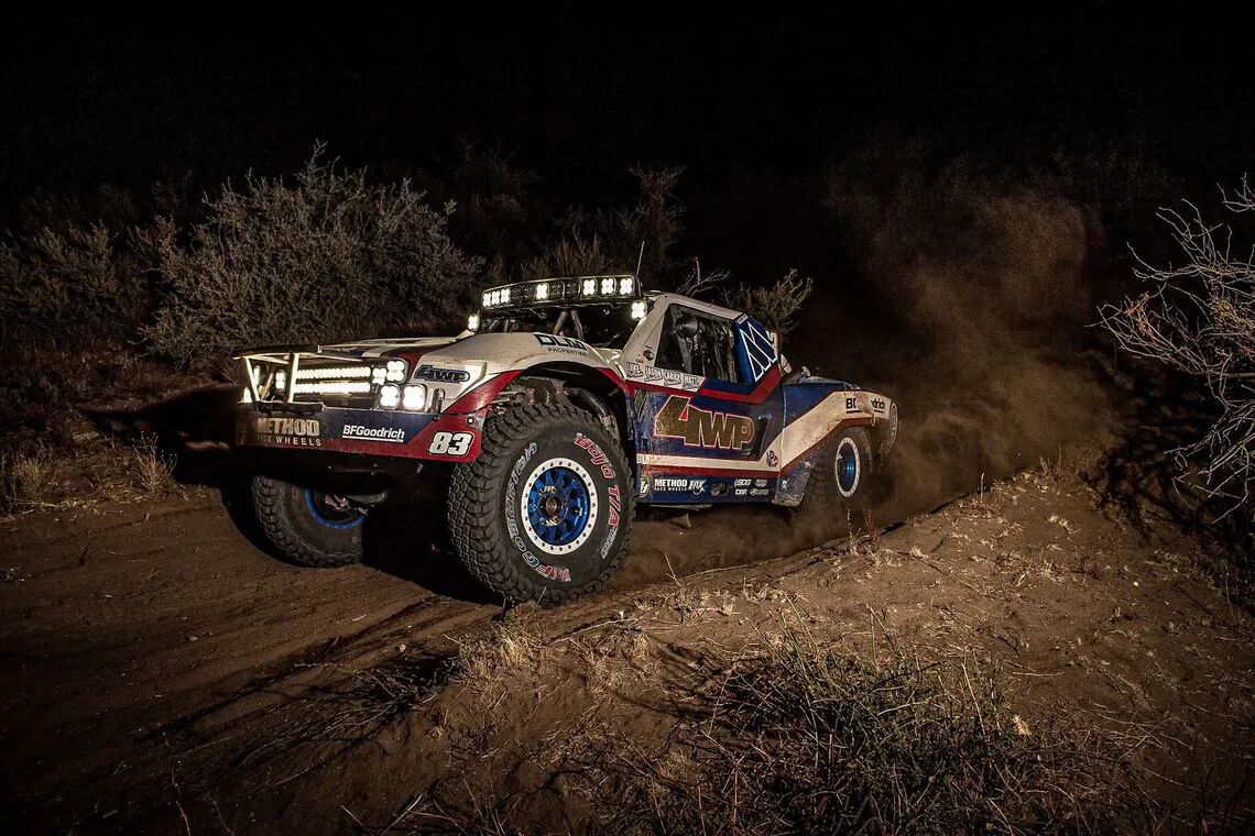 Terrängstävlingslastbil nummer 83, i rött, vitt och blått, med BFGoodrich-logon, kör genom natten över dammig terräng med sina strålkastare påslagna