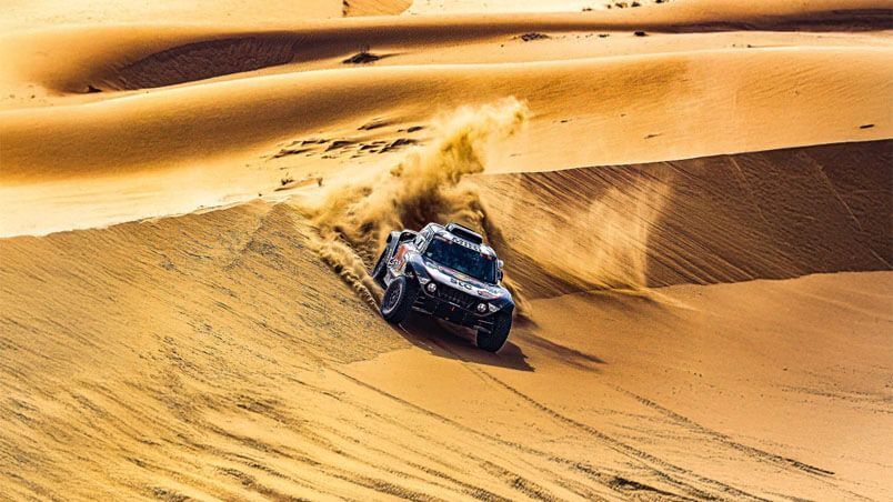 Auto da rally fuoristrada che attraversa una duna di sabbia nel deserto, sollevando una nuvola di polvere
