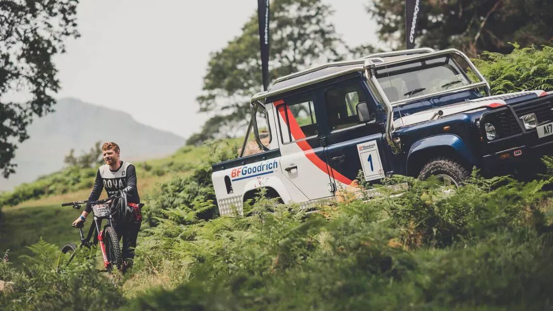 Ciclista con bicicleta de montaña junto a un 4x4 BFGoodrich en un exuberante paisaje natural con colinas y árboles de fondo