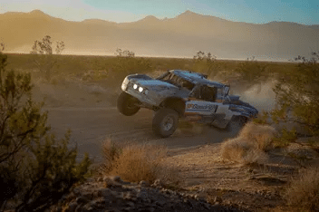 Terrängtävlingslastbil river upp damm när den kör genom ett ökenlandskap i solnedgången, med berg i bakgrunden