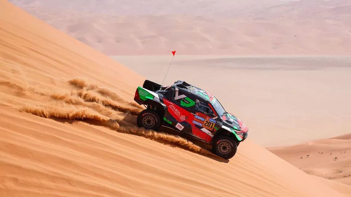 Terrängbil kör nedför en sanddyn i öknen under Dakar-rallyt, med en röd flagga på antennen och sandberg i bakgrunden