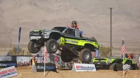 Camión de carreras en medio de un espectacular salto durante la carrera Mint 400, con banderas americanas y el logotipo de BFGoodrich al fondo