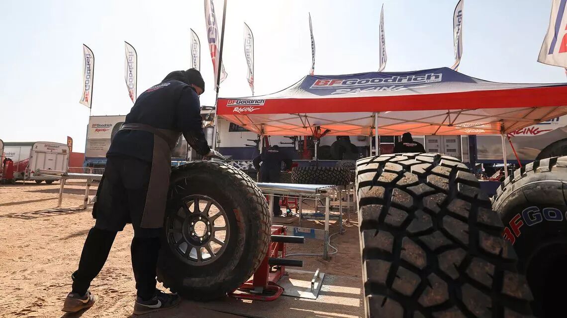 Mekaniker arbetar på ett stort offroad-däck i ett av BFGoodrichs tävlingstält i öknen, med flera däck och flaggor i bakgrunden