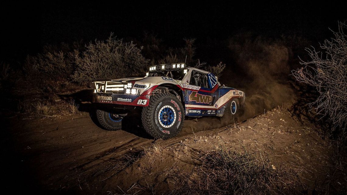 Camion da corsa fuoristrada numero 83, rosso, bianco e blu con il logo BFGoodrich guidato di notte su un terreno polveroso con i fari accesi.