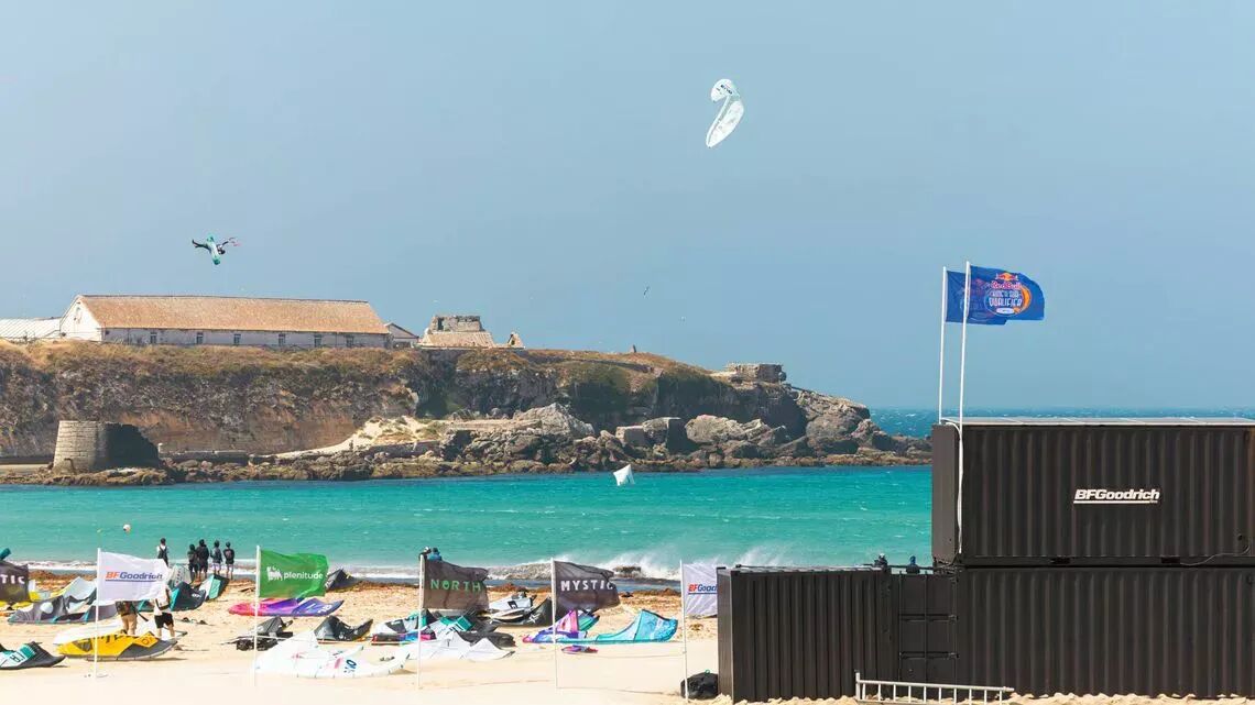 Kitesurfare flyger över havet under Red Bull King of the Air-tävlingen i Tarifa, med eventflaggor och tillfälliga konstruktioner på stranden i förgrunden