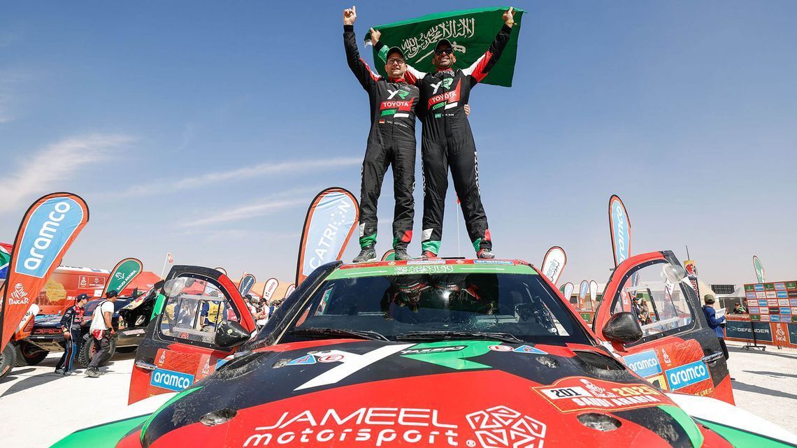 Piloti di rally che festeggiano la vittoria in piedi su un’auto rossa e verde, sventolando una bandiera a un evento automobilistico