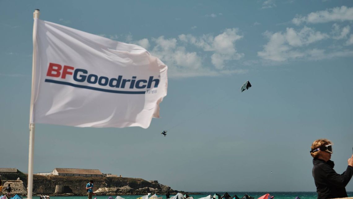 Drapeau BFGoodrich Tires flottant sur une plage avec une personne en kitesurf en arrière-plan, une autre personne debout à droite, ciel partiellement nuageux et bâtiments visibles au loin